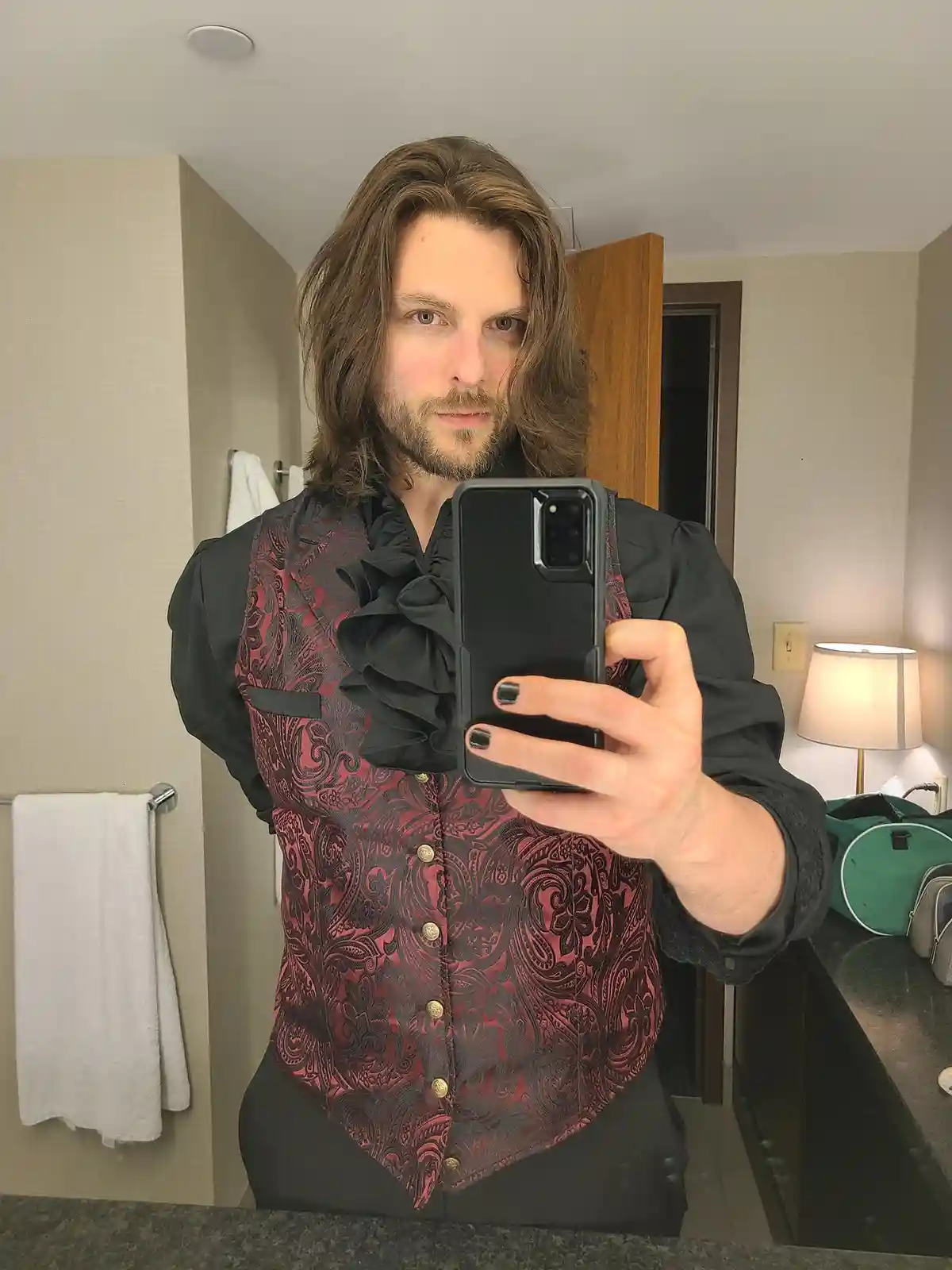 A man with long brown hair and a beard takes a mirror selfie in a hotel bathroom, wearing a black shirt with a large ruffled…