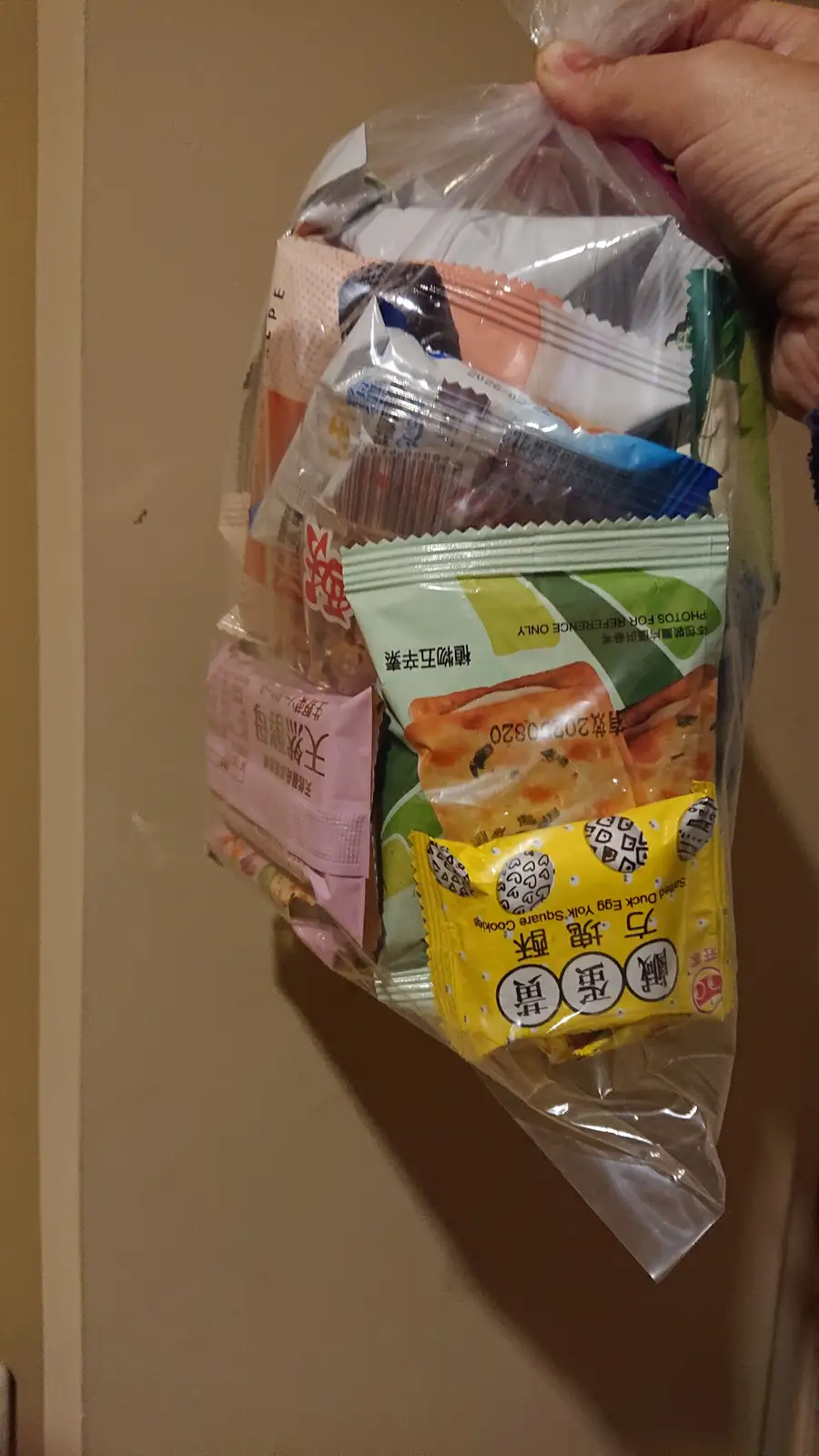 A hand holds a clear plastic bag overflowing with various small, individually wrapped Asian snacks, including a yellow…