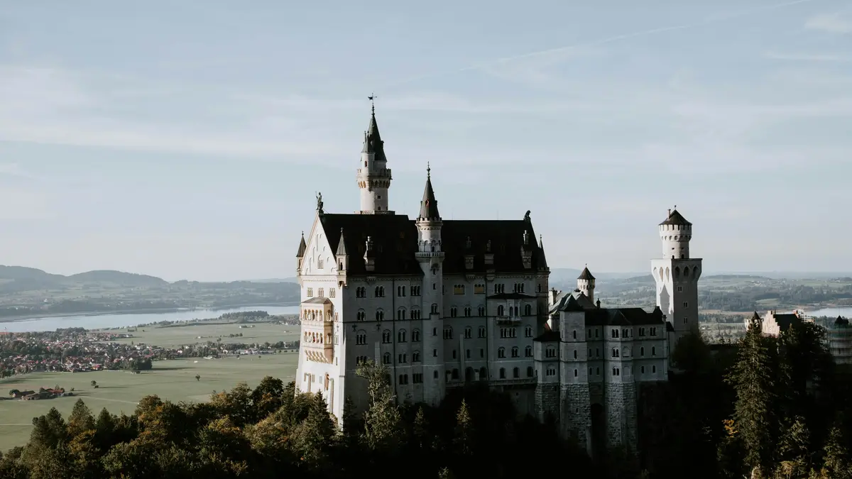 Towering majestically above the verdant valley, Neuschwanstein Castle's turrets and spires reach for the sky, surrounded by…