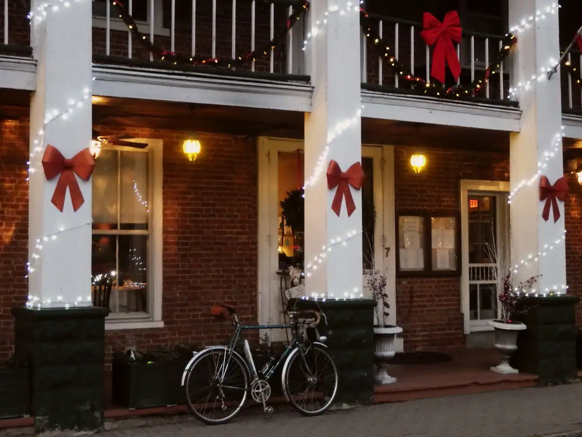A bicycle is parked on the sidewalk in front of a brick building decorated with festive red bows and string lights on white…