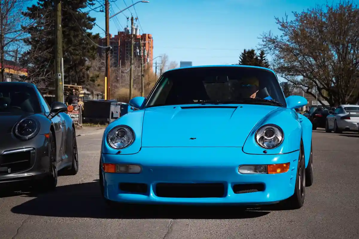 A bright blue Porsche 911 is parked on a street with other cars, including a gray Porsche 911 beside it. A man wearing a…