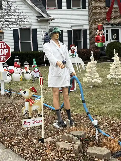A mannequin dressed in a white robe, green hat, and socks stands in a yard holding a beer can and a blue vacuum hose…