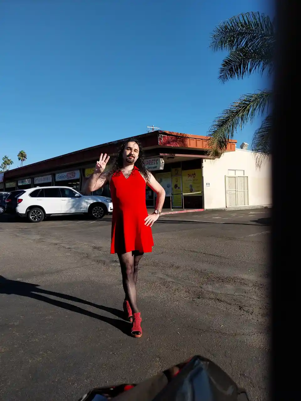 A man with long dark hair and a beard stands in a parking lot in front of a strip mall, wearing a bright red dress, black…