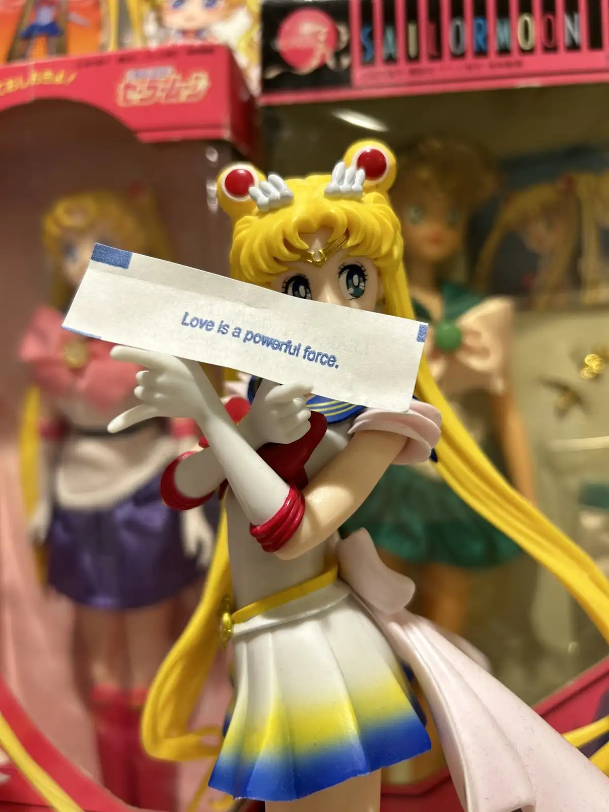 A close-up of a Sailor Moon anime figurine holding a small paper slip that reads 'Love is a powerful force.' The figurine is…