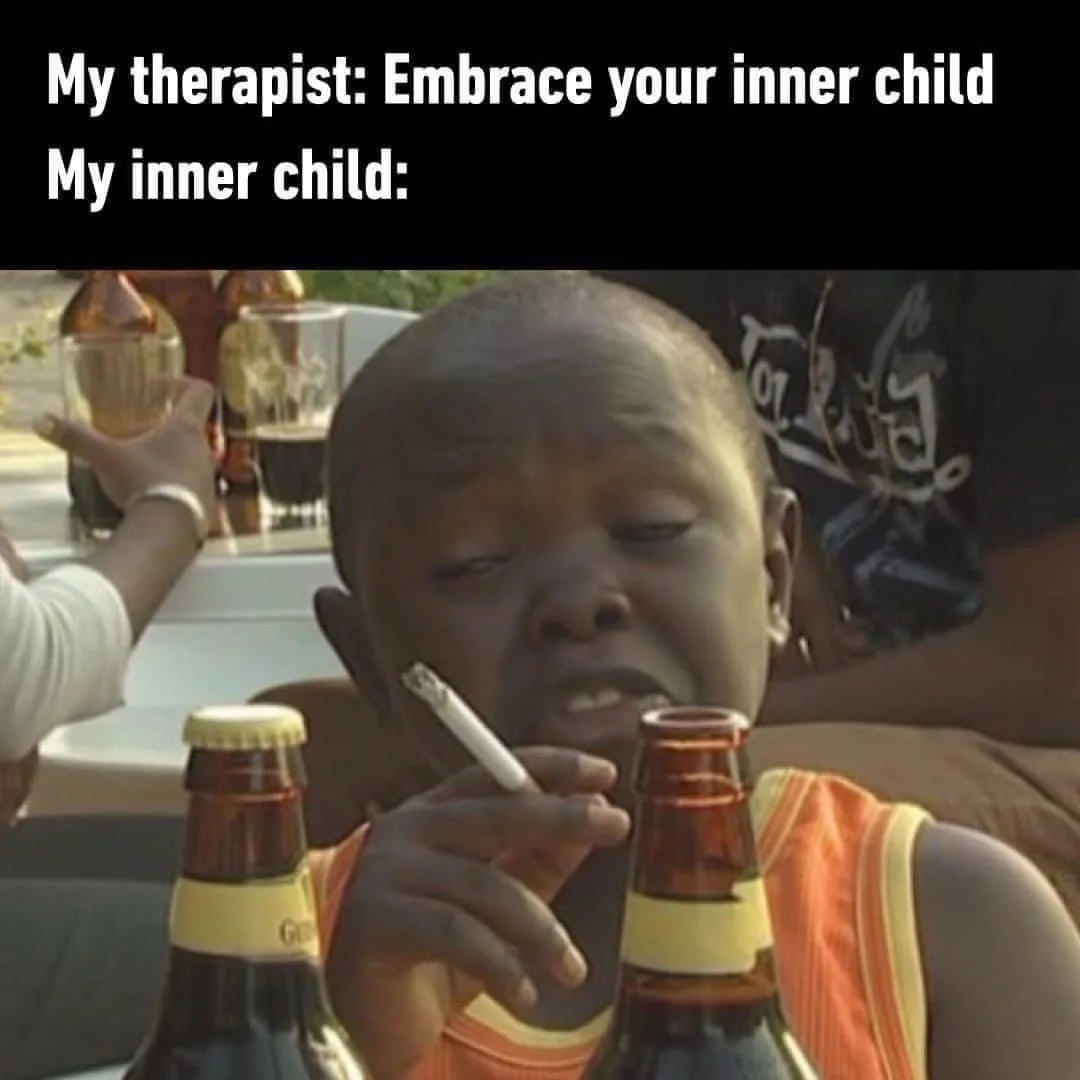 A young child with a shaved head, wearing an orange and yellow tank top, is seated at a table holding a lit cigarette…