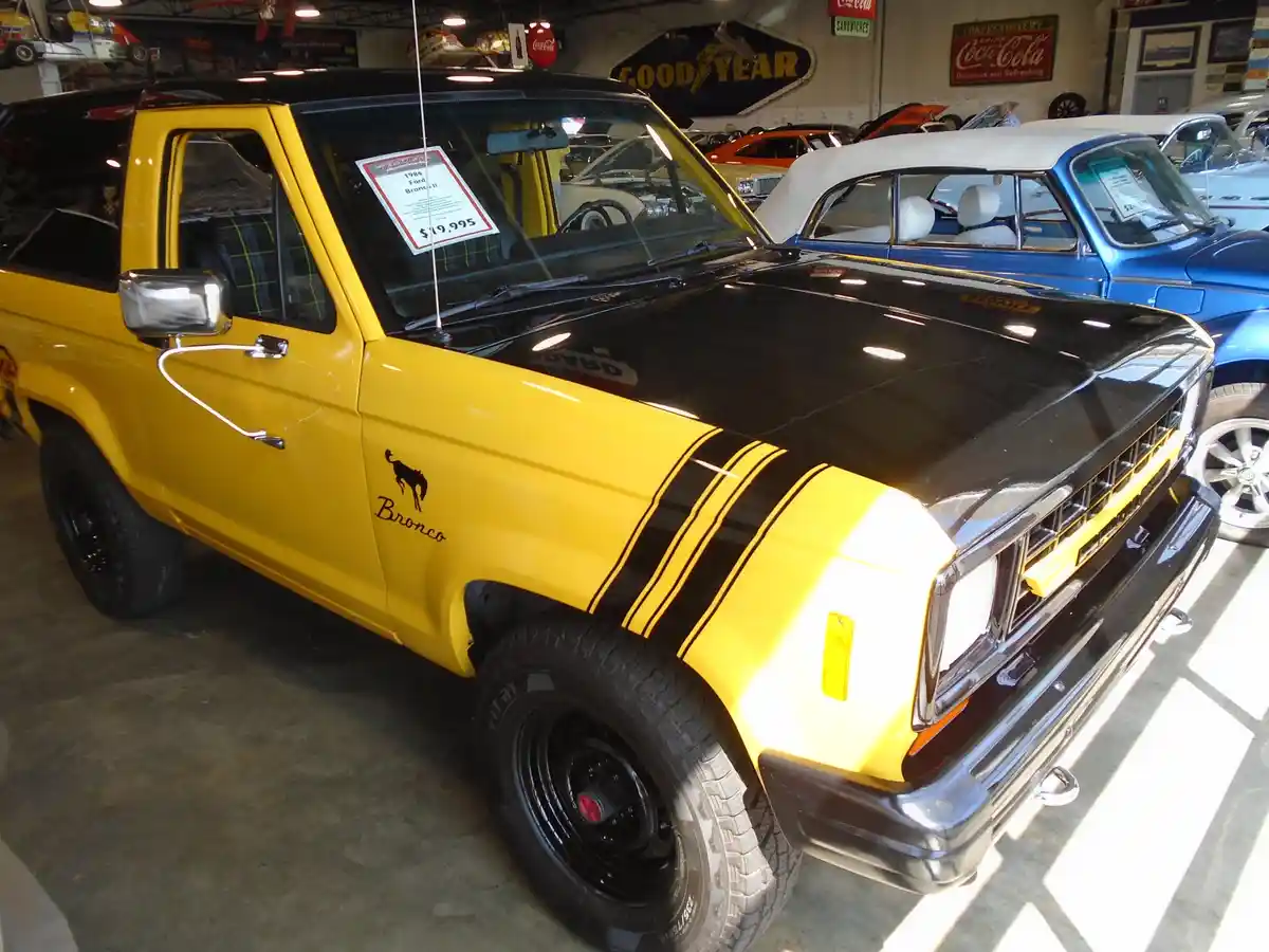 A yellow and black 1984 Ford Bronco II is displayed in a car showroom, with a price tag of $19,995 visible on the…