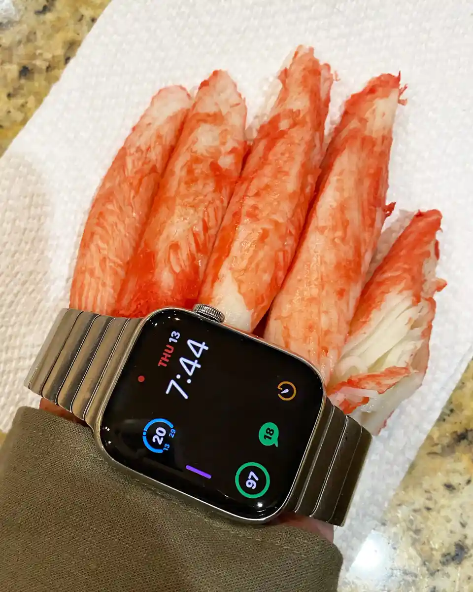A smartwatch with a metallic band is positioned on a white paper towel, surrounded by five imitation crab sticks arranged…