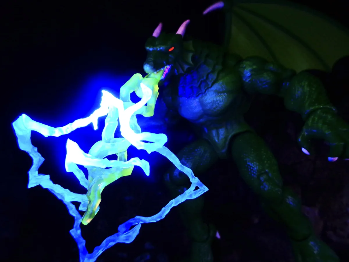 A green plastic dragon action figure with glowing red eyes and purple horns is shown in a dark setting, emitting a bright…