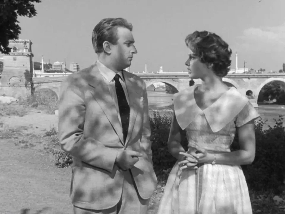 a black-and-white still from a classic film shows a man in a suit and a woman in a dress standing outdoors near a bridge…