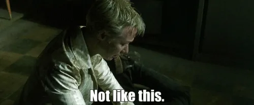 A man with blond hair, wearing a white shirt, is sitting on the floor in a dimly lit room, looking down with a defeated…