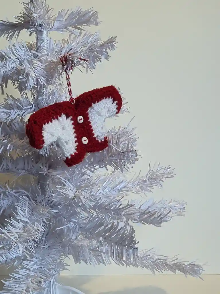 A red and white crocheted ornament shaped like a stylized fox or cat head hangs from a white artificial Christmas tree. The…