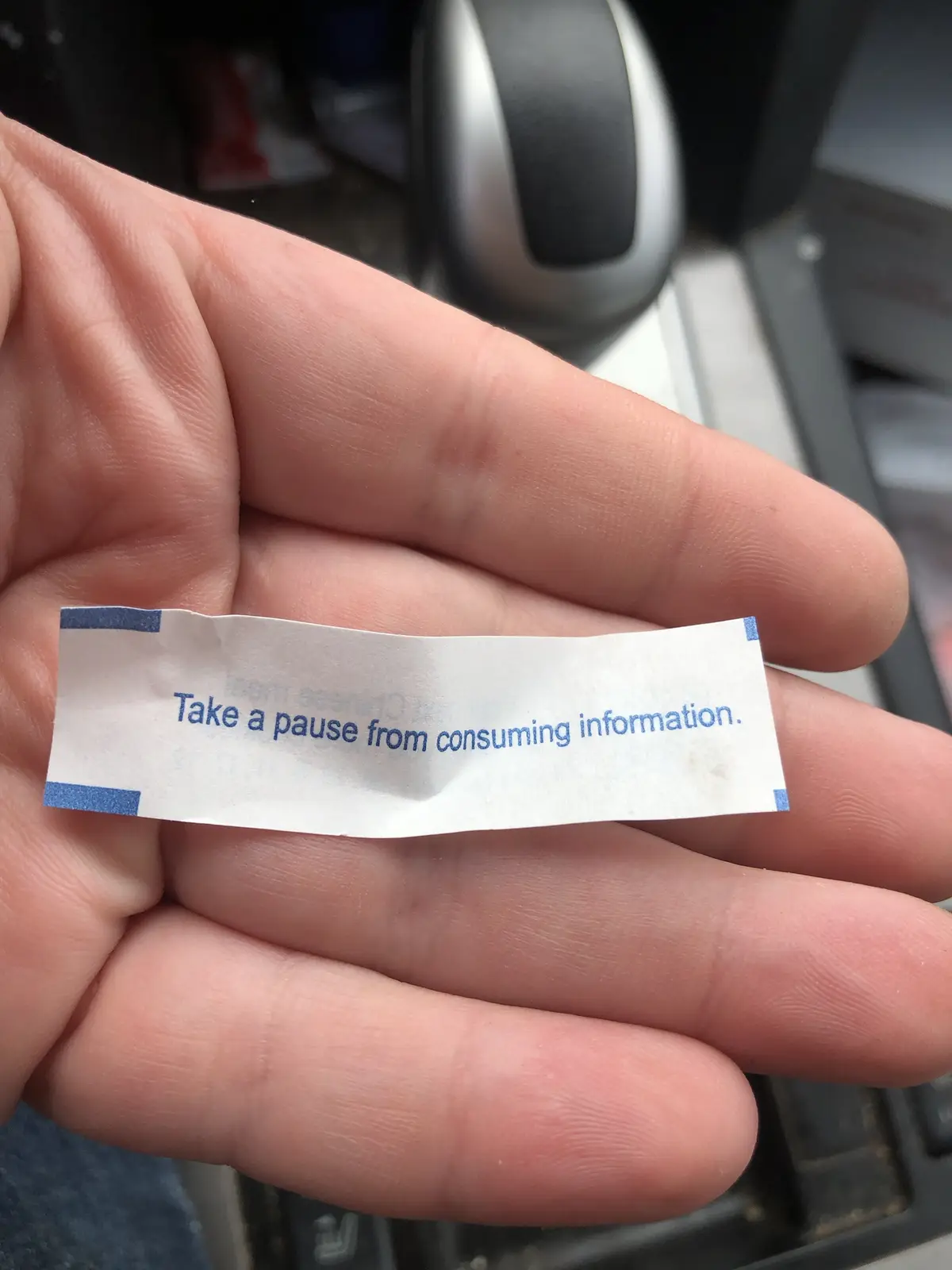 A person's hand holds a small, white fortune cookie slip with blue text that reads 'Take a pause from consuming…