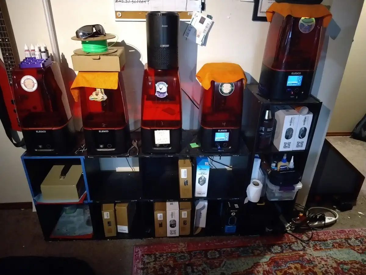 A row of five red and black Elegoo resin 3D printers sits on a black shelving unit, each labeled with a number and some…