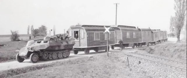 A black-and-white historical photo shows a military half-track vehicle pulling a passenger train car across a rural road…