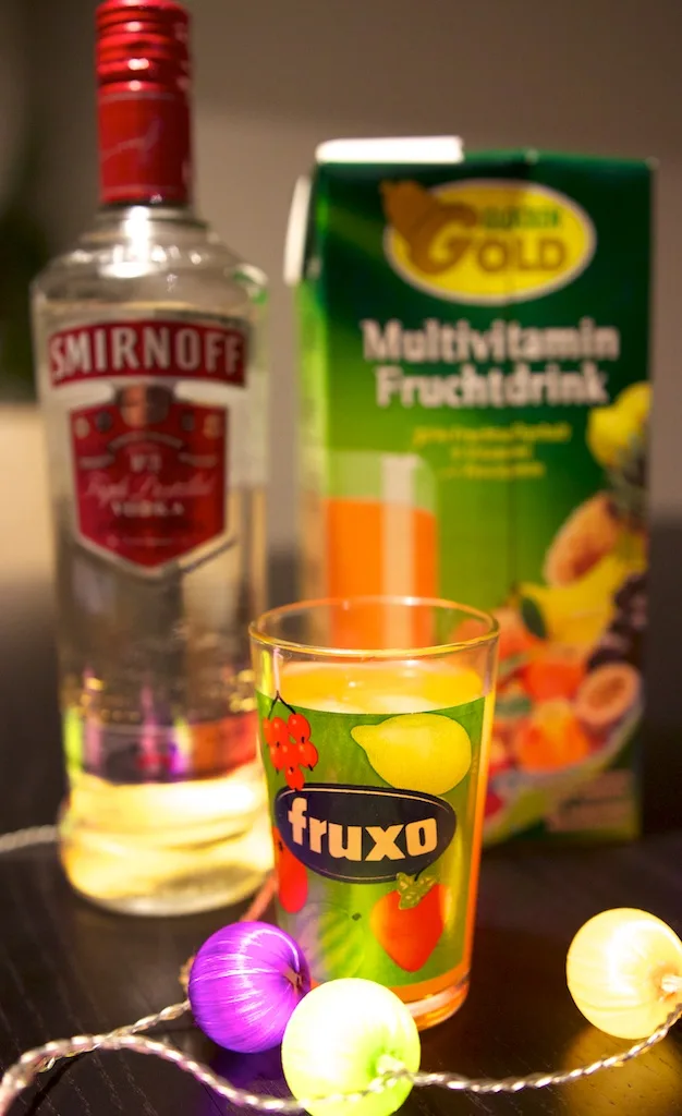 A glass labeled 'fruxo' filled with a yellow drink sits in front of a bottle of Smirnoff vodka and a box of Golden Gold…