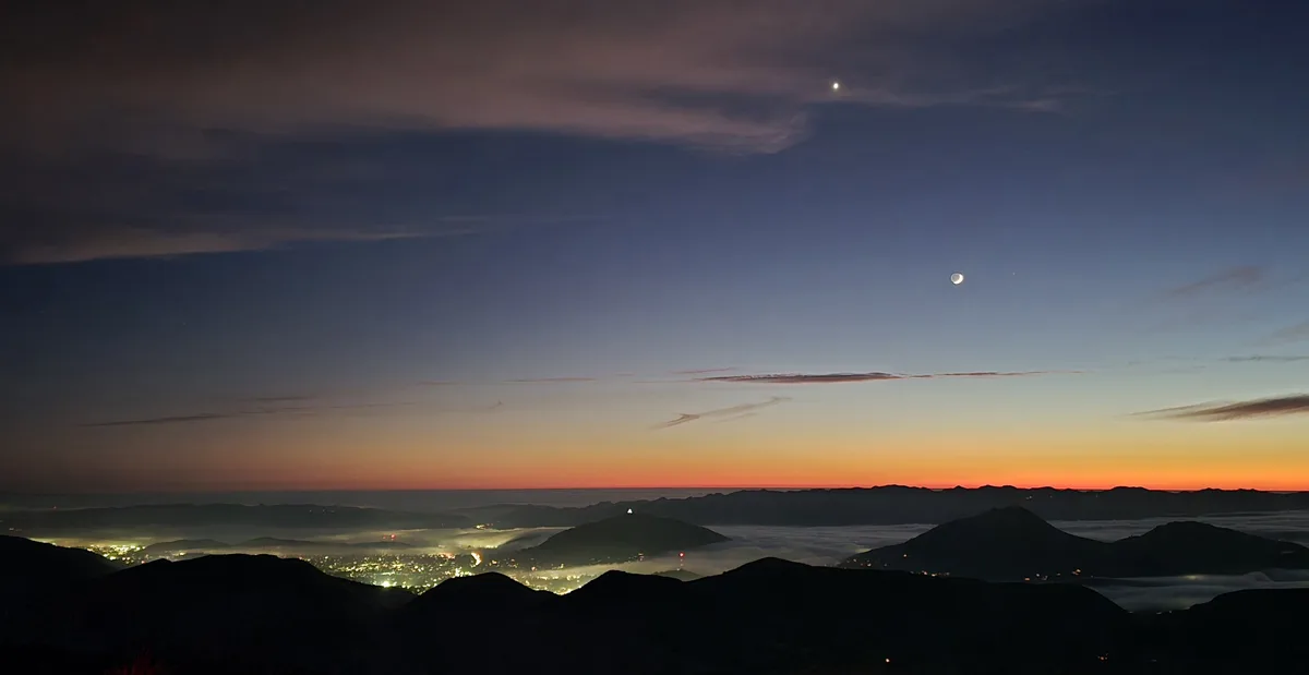 A panoramic twilight photo shows a crescent moon and a bright star in the sky above a valley filled with fog and city lights…