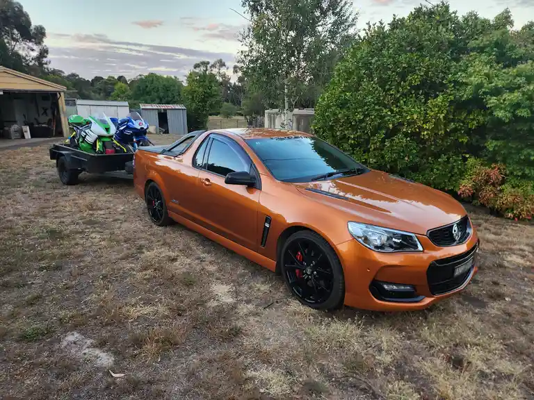 An orange Holden Ute pickup truck with black wheels and red brake calipers is parked on dry grass, towing a trailer carrying…