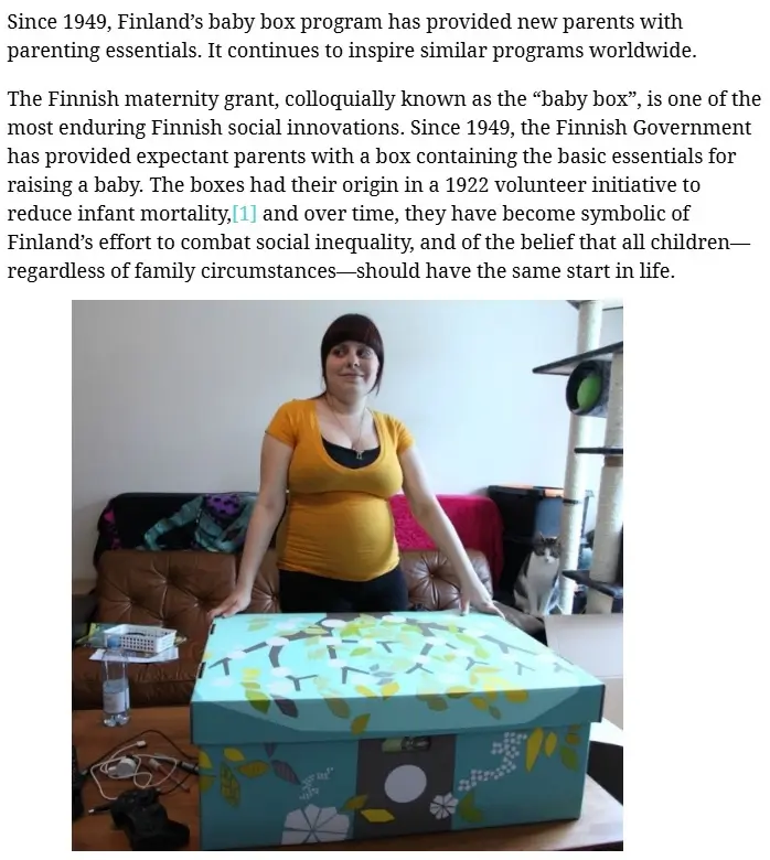 A pregnant woman in a yellow shirt stands behind a large, decorated baby box placed on a table. The box has a light blue lid…