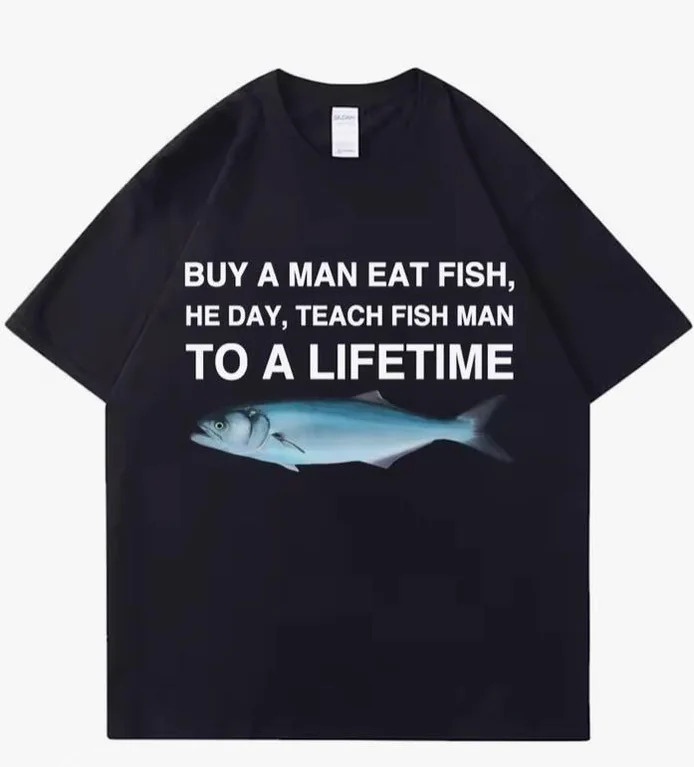 A black t-shirt displays white text reading 'BUY A MAN EAT FISH, HE DAY, TEACH FISH MAN TO A LIFETIME' above a graphic of a…