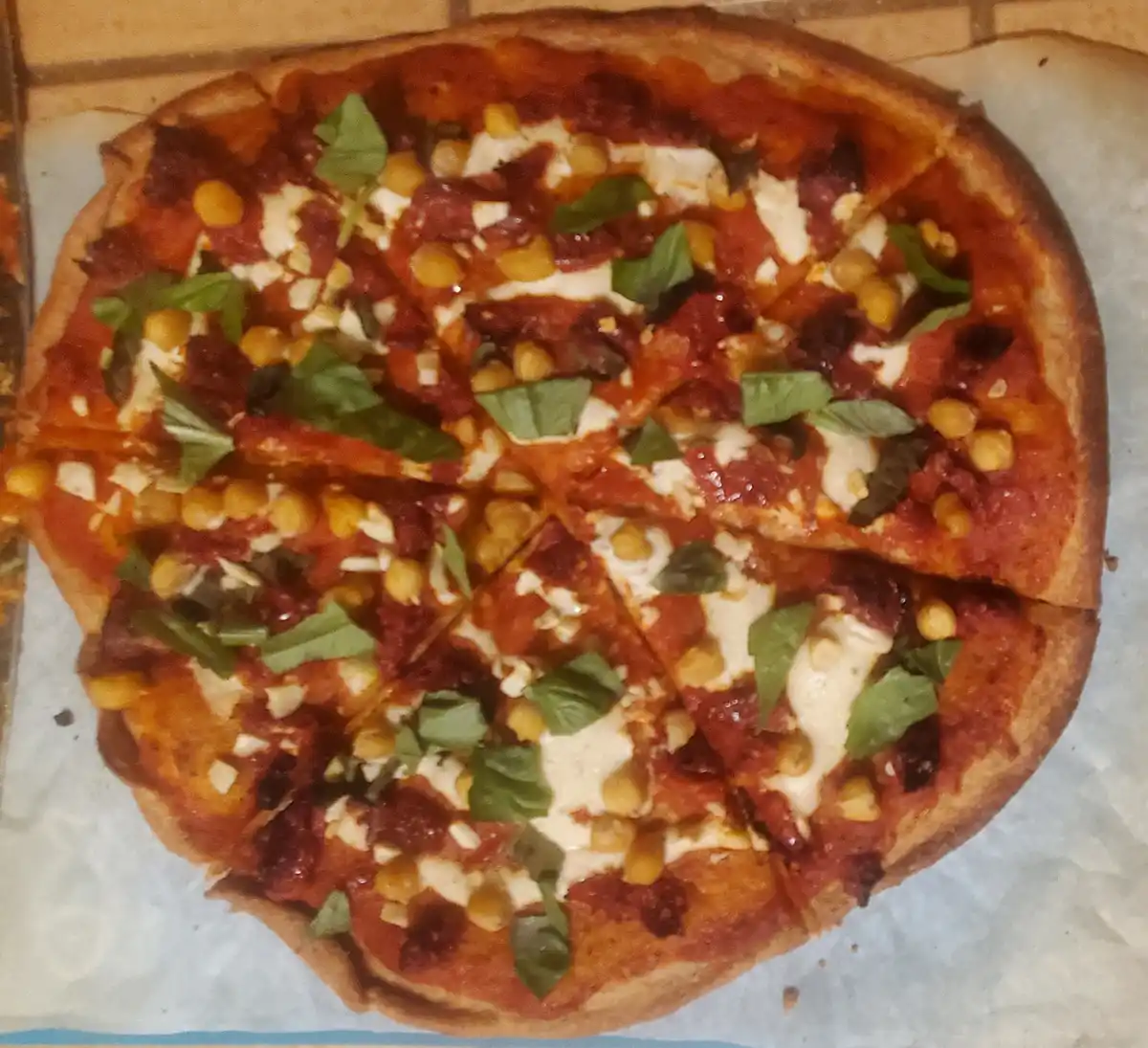 A close-up photo of a round, sliced pizza with tomato sauce, melted cheese, corn kernels, and fresh basil leaves on a…