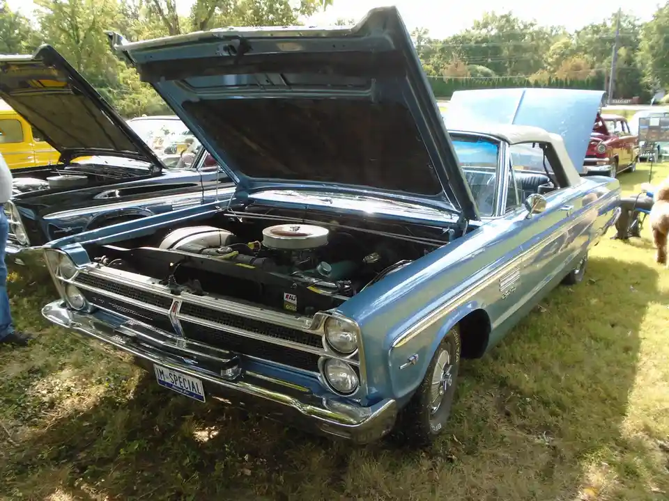 A light blue classic American convertible with its hood open, revealing a chrome engine, parked on grass at a car show with…