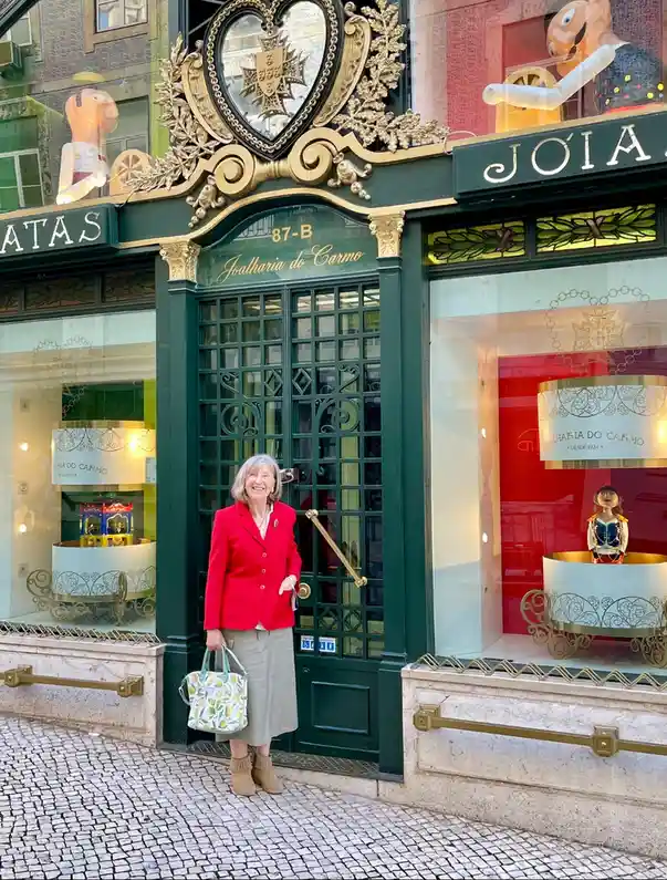 A woman in a red blazer and beige skirt stands smiling in front of a green ornate door of a jewelry store named 'Joalharia…