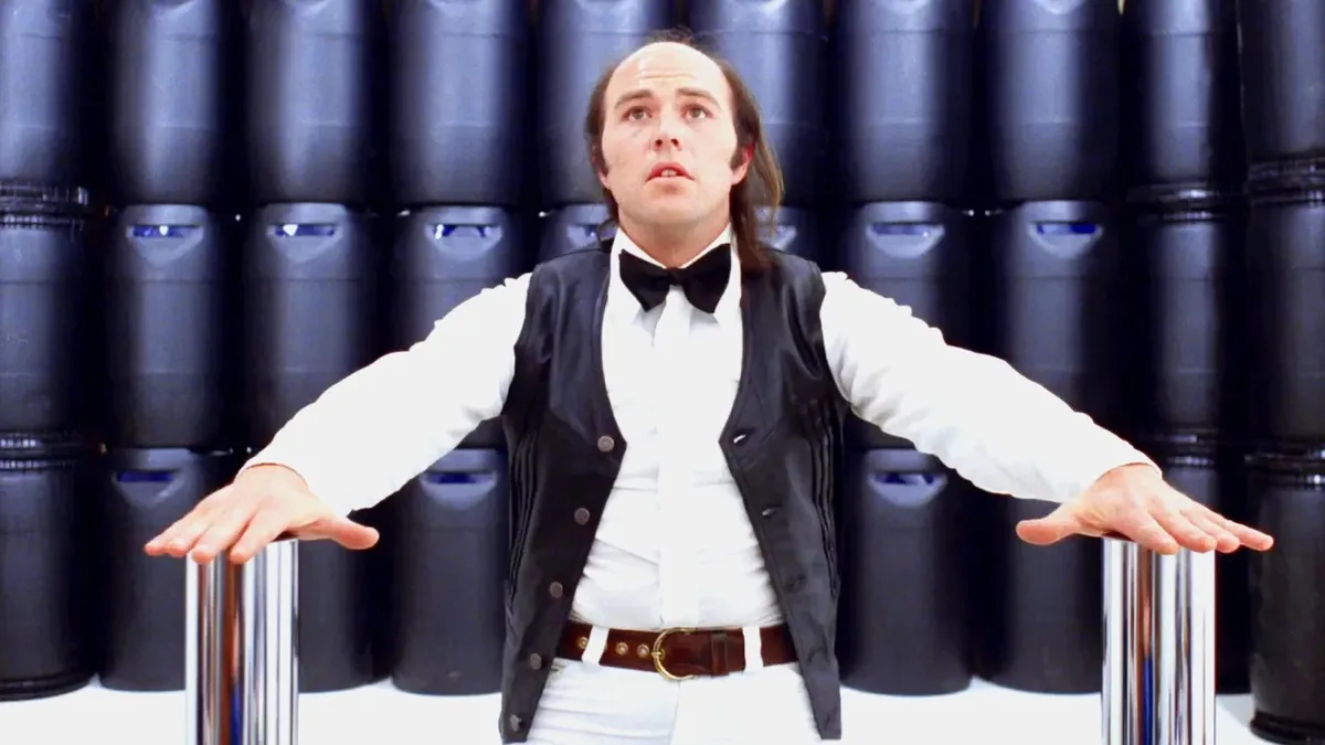 a man in a white shirt, black vest, and bowtie stands with arms outstretched, palms up, looking upward in confusion or…