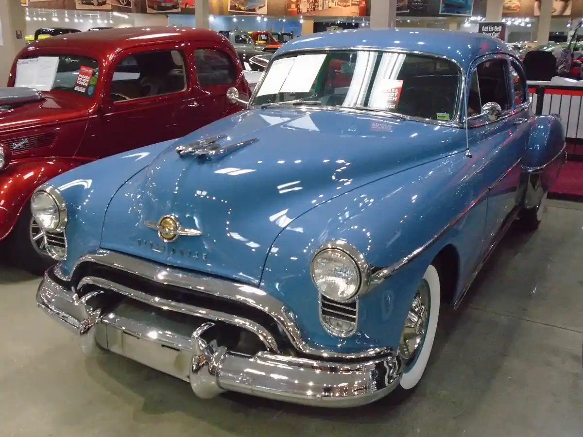 A shiny blue 1950s Oldsmobile classic car is displayed in a museum or showroom, with a red vintage car visible behind it…