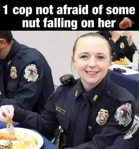 A smiling female police officer in uniform sits at a table eating food, with a male officer visible beside her. The image…