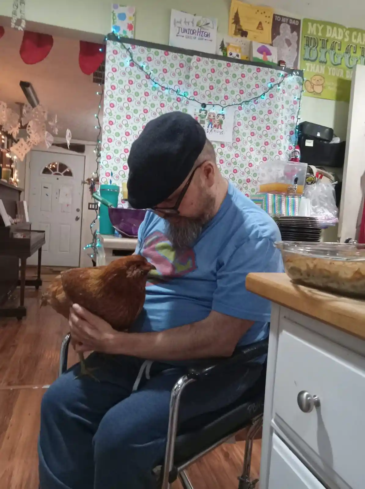 A man with a beard, glasses, and a black beret sits in a wheelchair in a decorated kitchen, gently holding a brown chicken…