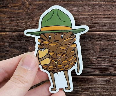 A hand holds a die-cut sticker of an anthropomorphic pinecone wearing a green ranger hat with a yellow band, holding a…