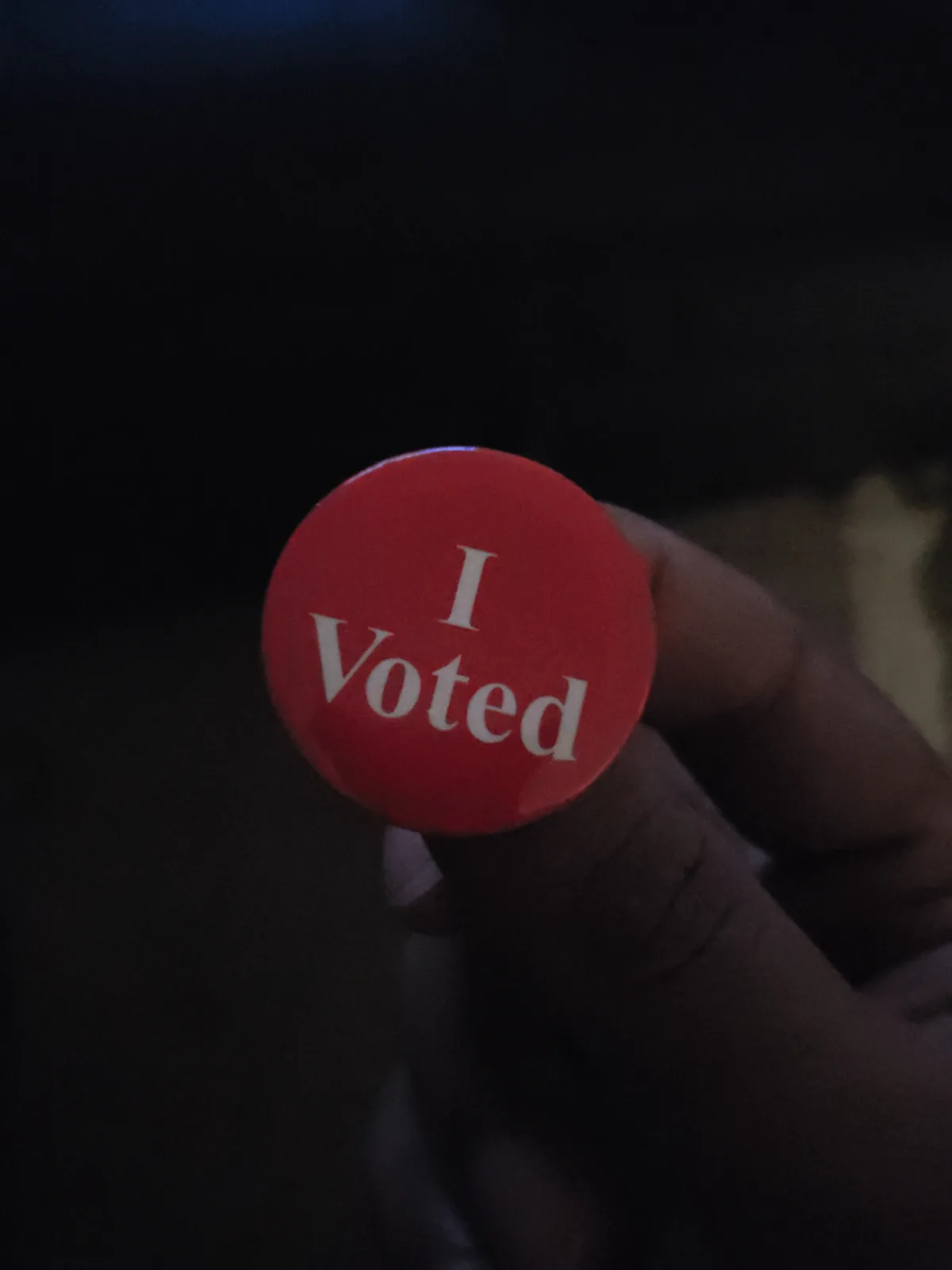 A close-up photo shows a hand holding a small, round, red button with the white text 'I Voted' printed on it. The background…