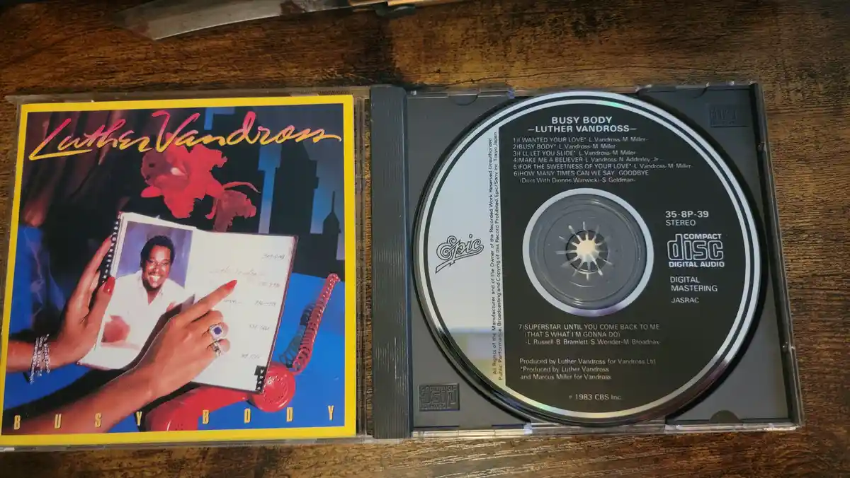 An open CD case displays Luther Vandross's 1983 album 'Busy Body', showing the colorful cover art with a woman holding a…