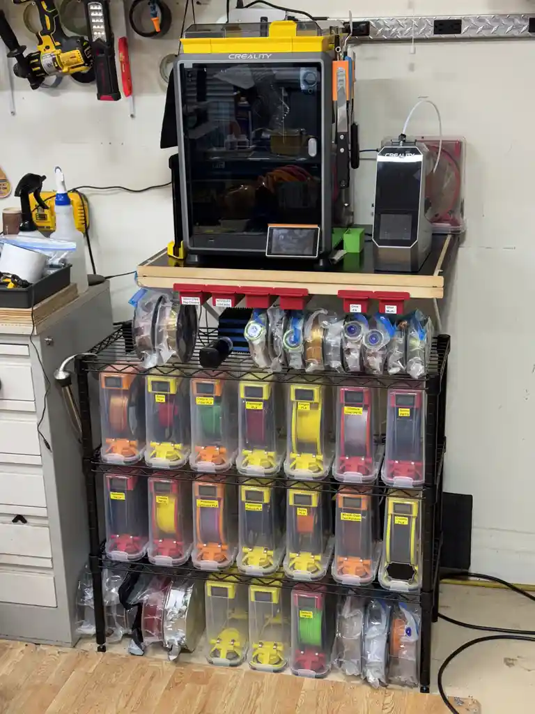 A well-organized 3d printing station features a Creality printer on a shelf above a black metal rack holding dozens of…