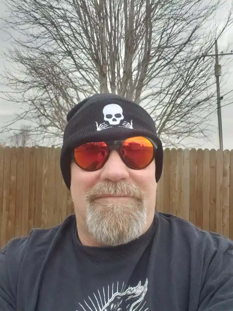 A man with a gray beard and mustache wears a black beanie with a white skull and crossbones design and large orange-tinted…