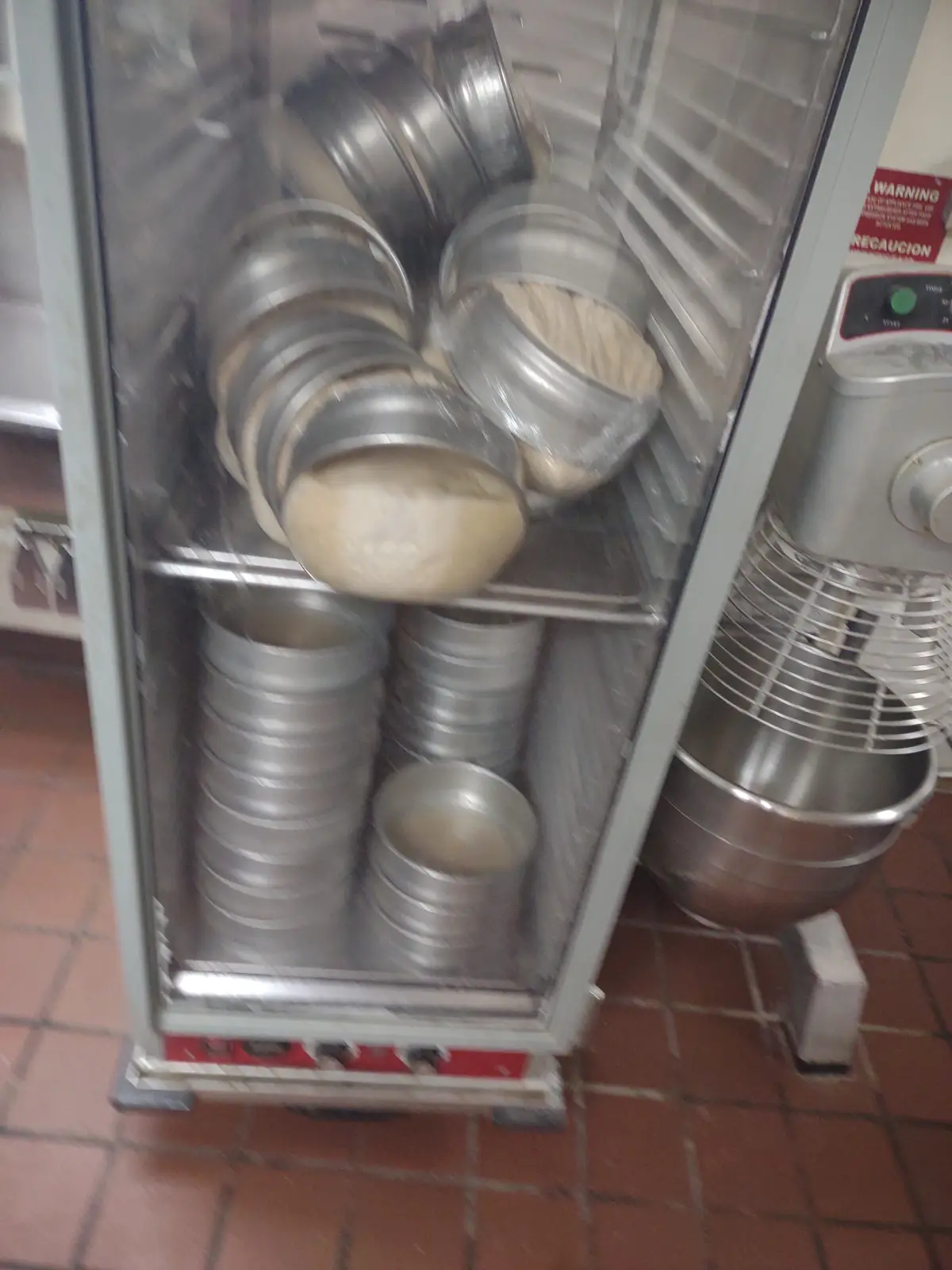 A commercial proofing cabinet filled with metal baking pans, some containing risen dough balls, sits in a bakery kitchen…