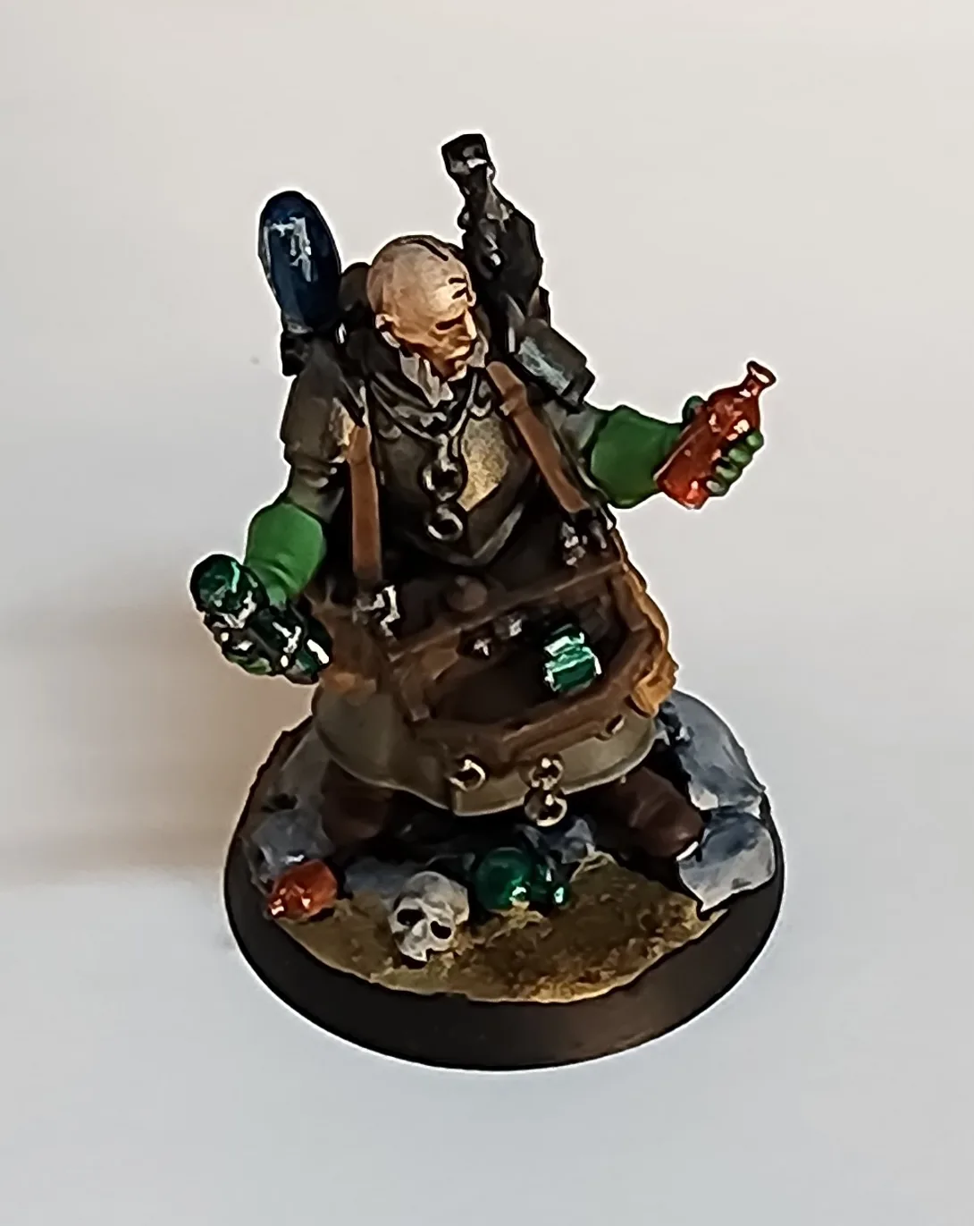 A painted miniature figurine of a bald, bearded man in adventurer's gear, holding a red bottle and a flask, with a backpack…