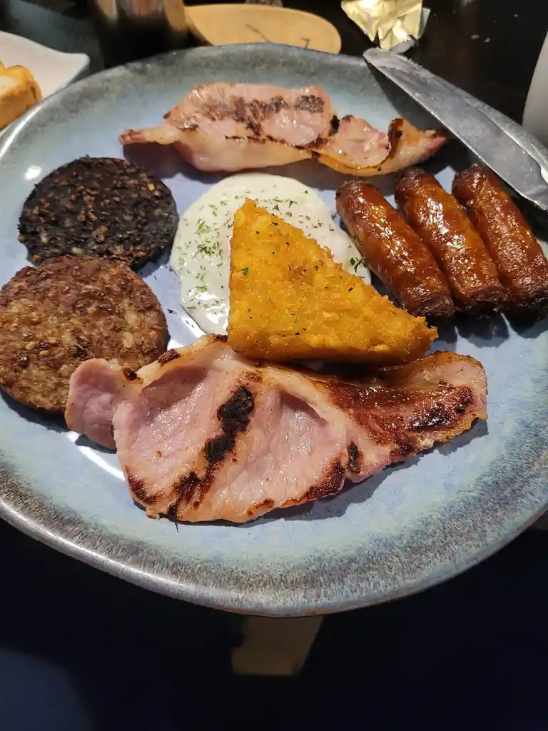 A close-up photo of a full English breakfast on a blue ceramic plate, featuring grilled bacon, ham, three sausages, a fried…
