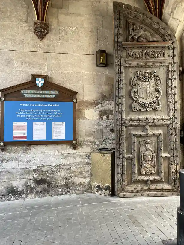 A photo of a historic stone building entrance with a large, ornate wooden door featuring carvings and a coat of arms. To the…