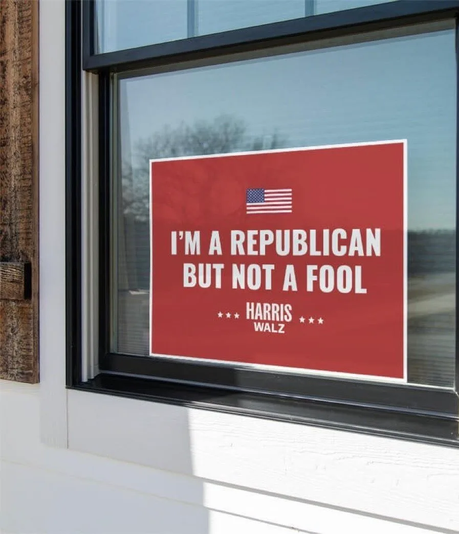 A red political sign is affixed to a window of a white-sided house, featuring a small American flag at the top, followed by…