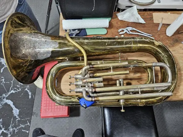 A brass sousaphone rests on a wooden workbench surrounded by tools like wrenches and paper towels. A person's legs and feet…