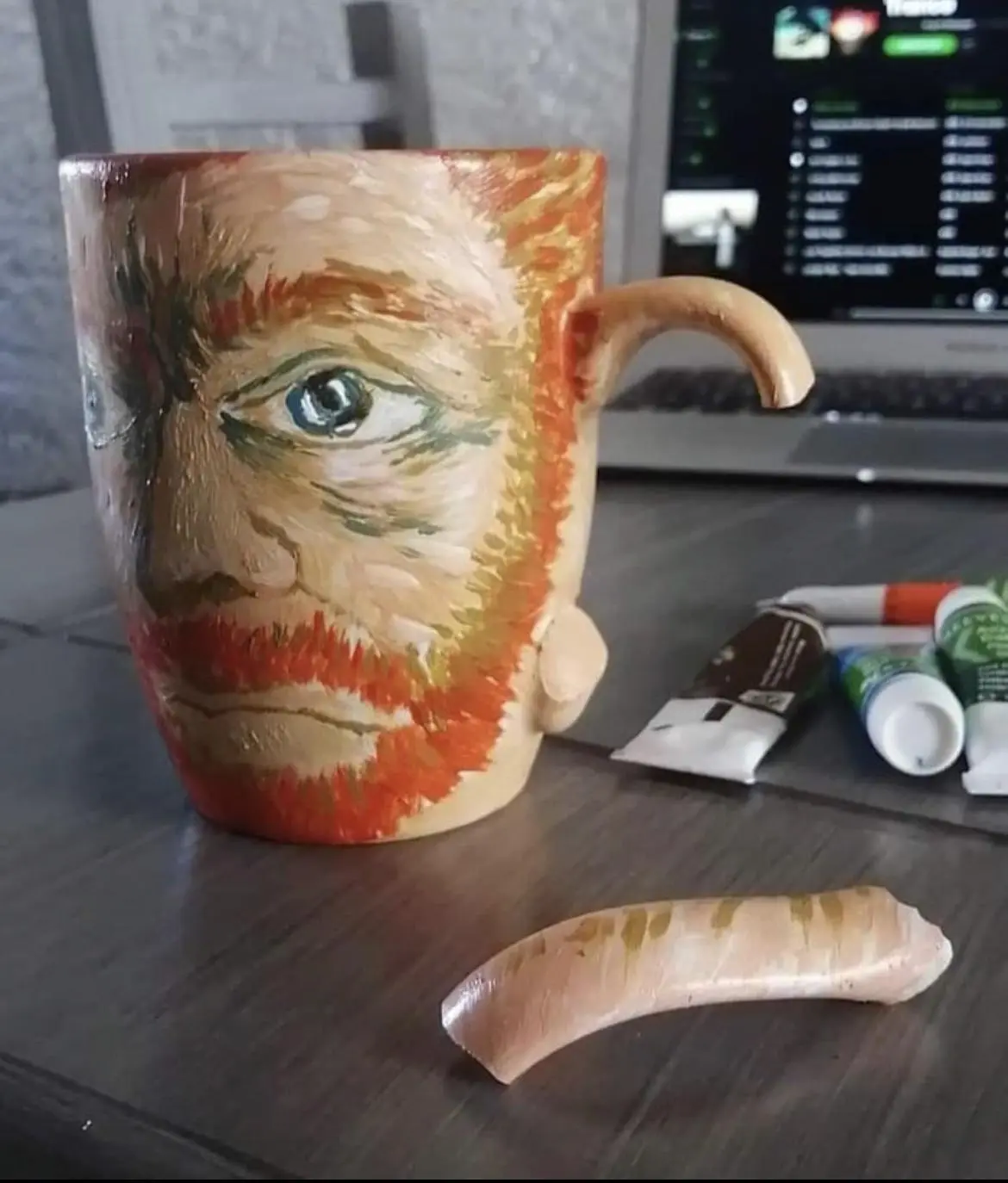 A ceramic mug painted with a portrait of Vincent van Gogh, featuring his distinctive blue eye and red beard, sits on a…
