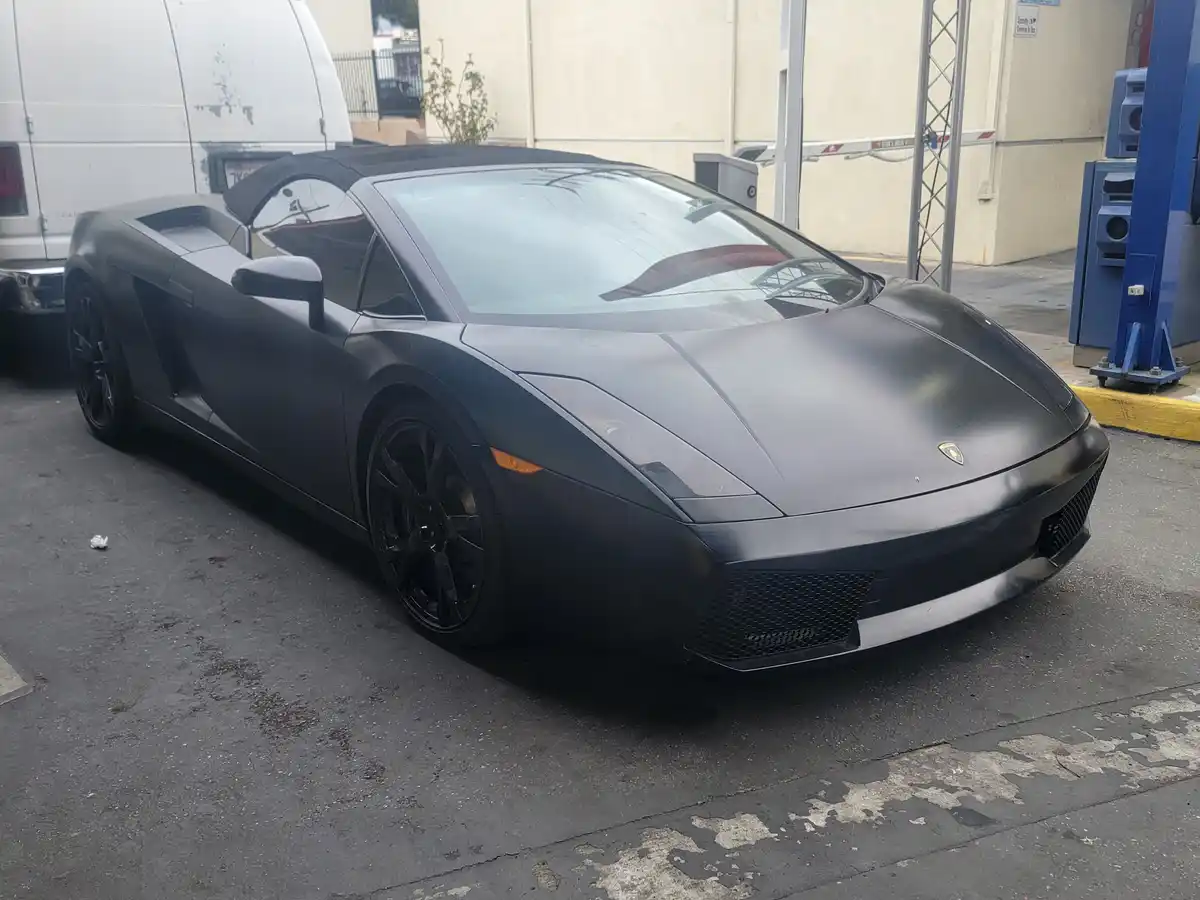 a matte black lamborghini gallardo convertible is parked in a worn asphalt lot next to a white van and a blue car lift, with…