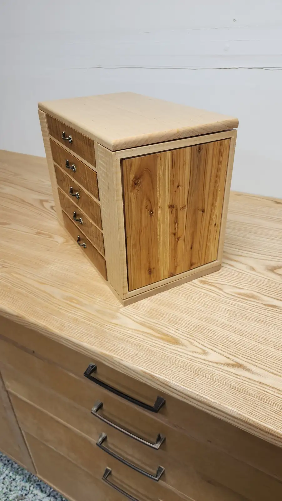 A small, handmade wooden box with four drawers and a solid wood front panel sits on a larger wooden dresser. The box has…