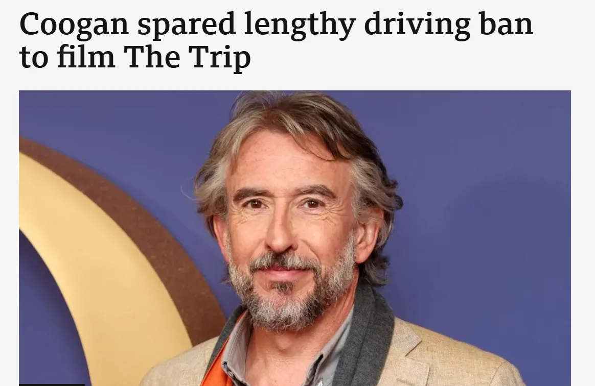 A headshot of actor Steve Coogan, who has gray hair and a beard, wearing a beige blazer over an orange shirt and gray scarf…