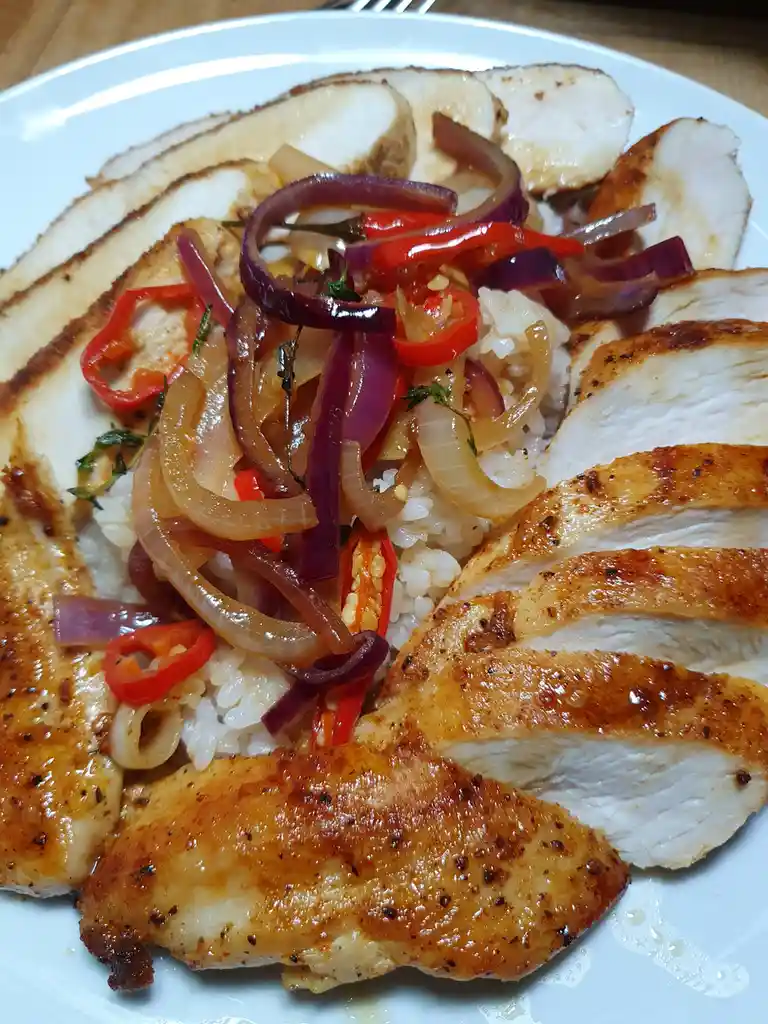 A close-up photo of a plated meal featuring sliced grilled chicken breast arranged around a bed of white rice, topped with…