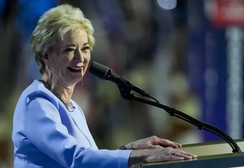 A woman with short blonde hair, wearing a light blue suit and jewelry, stands at a podium with a microphone, smiling broadly…