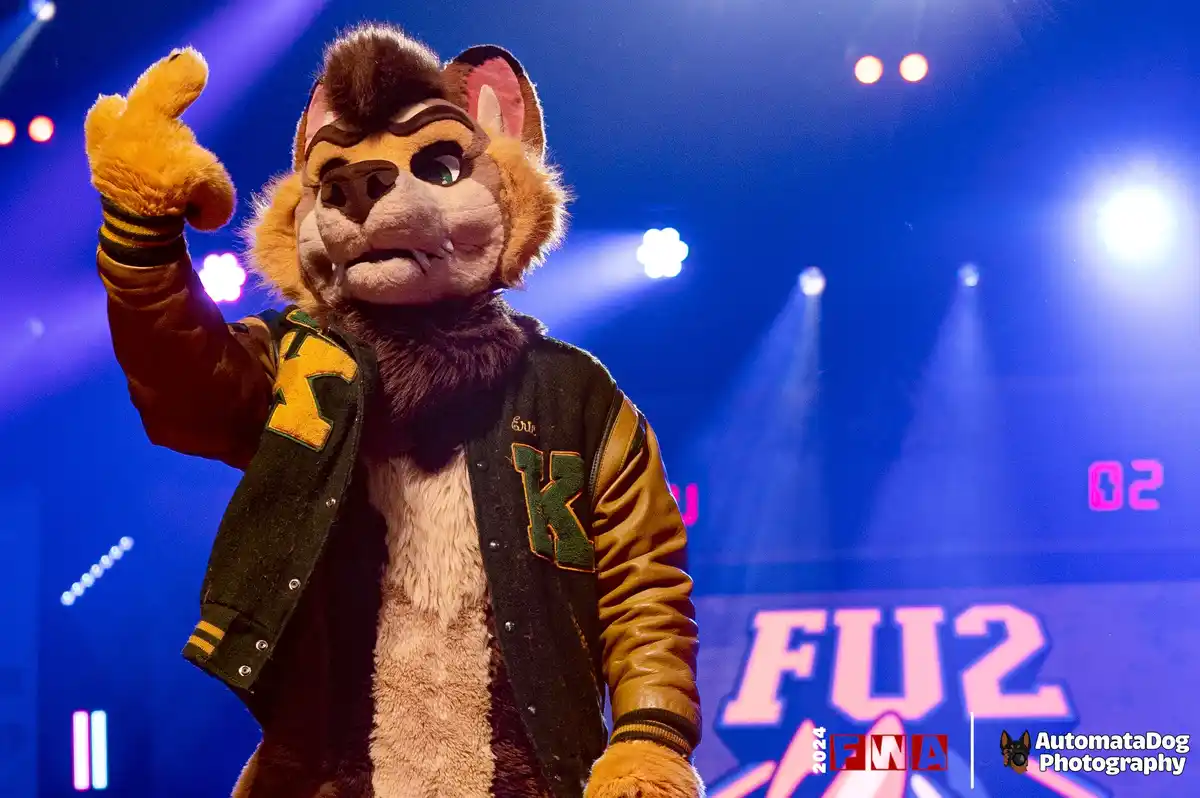 A person in a detailed fursuit of a stylized fox or wolf mascot, wearing a varsity jacket with a large 'K' on the chest and…