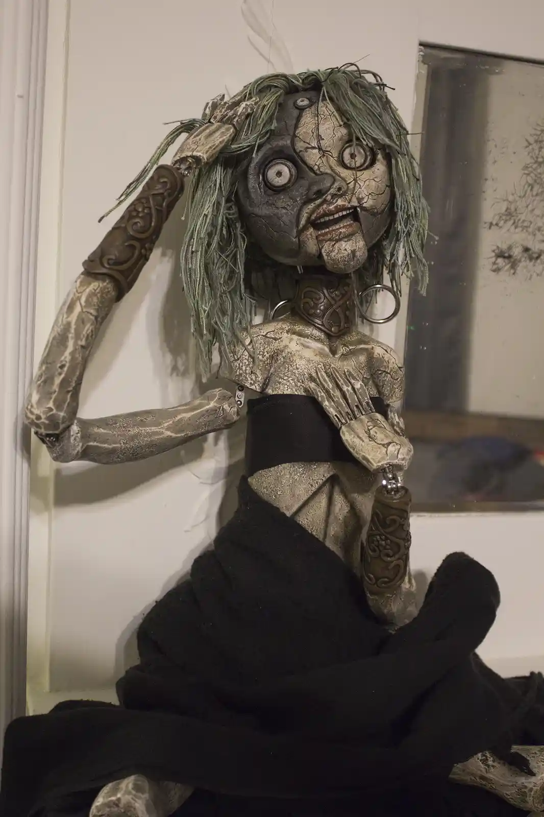 A highly detailed, unsettling doll with cracked, grayish skin, wild green yarn hair, and articulated limbs sits draped in…