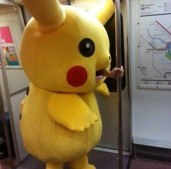 A person in a full-body yellow Pikachu costume stands inside a subway car, gripping a pole with one hand while their face is…